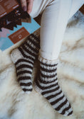 Load image into Gallery viewer, Woman sitting on sheepskin rug and wearing warm and cosy hand knitted pair of wool socks. Reading the Bamboshe care instructions