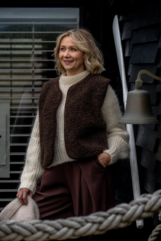 Woman wearing a Freya espresso brown merino wool vest with a cream knit jumper and brown trousers.
