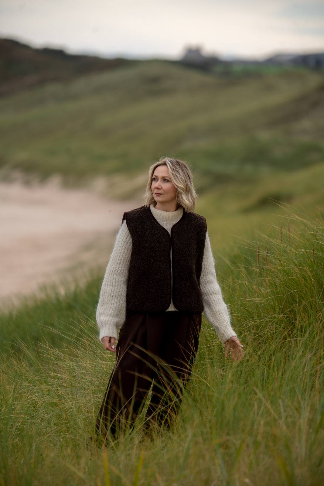 Freya espresso brown merino wool vest worn over a cream jumper on a walk through coastal grassland.