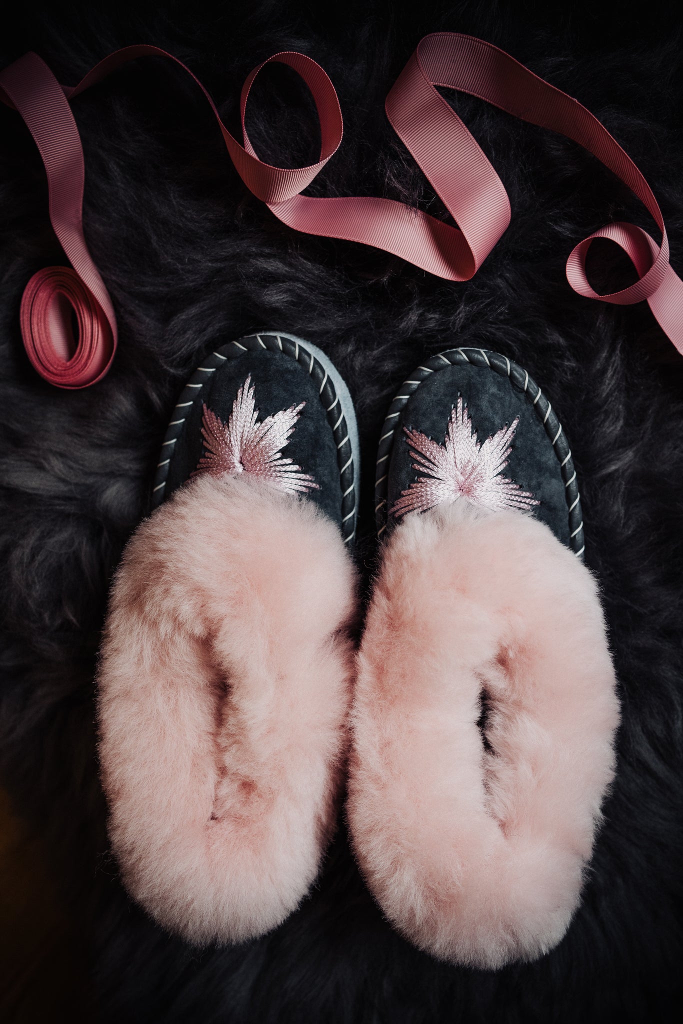  Pink candy sheepskin slippers with a pink rim and grey leather, exquisitely hand-embroidered with pink silk thread, resting on a fluffy grey sheepskin with pink ribbon decoration.