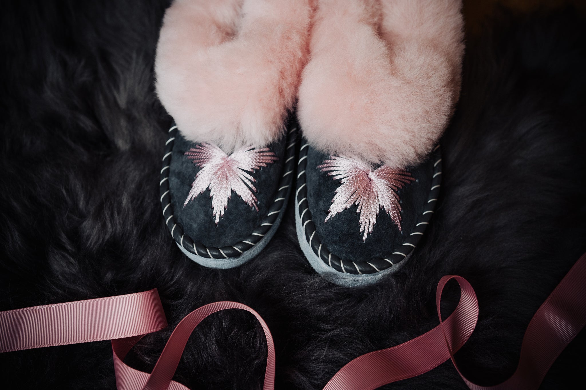 Pink candy sheepskin slippers with a pink rim and grey leather, exquisitely hand-embroidered with pink silk thread, resting on a fluffy grey sheepskin with pink ribbon decoration.