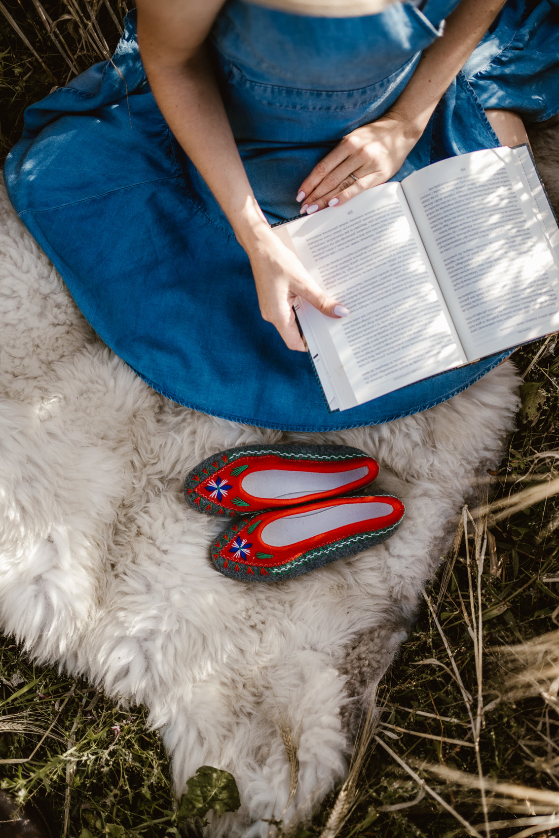 Woman sitting on soft fluffy sheepskin. She is reading book in nature feeling relax and cosy. In front of her are wool felt handmade folk, boho style slippers