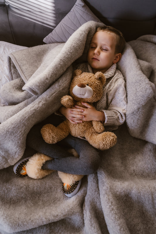 Sleeping Childe wrapped in Pure Merino Wool Blanket holding teddy bear. He is wearing sheepskin fur slippers feeling warm and cosy