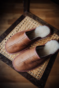 Load image into Gallery viewer, Soft men's leather slippers with cream wool lining, elegantly displayed on a nice chair.
