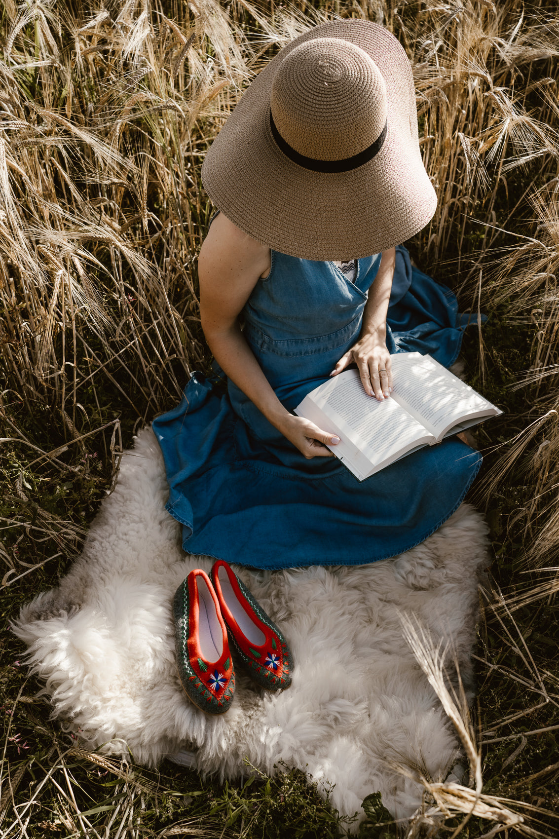 Woman sitting on soft fluffy sheepskin. She is reading book in nature feeling relax and cosy. In front of her are wool felt handmade folk, boho style slippers