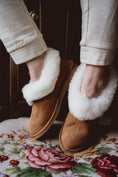 Load image into Gallery viewer, Alt text: Classic sheepskin slippers with white fur, worn by a woman and placed on a hand-embroidered chair.
