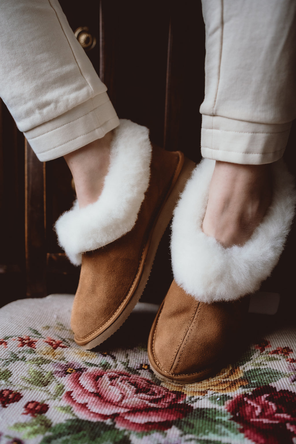 Alt text: Classic sheepskin slippers with white fur, worn by a woman and placed on a hand-embroidered chair.