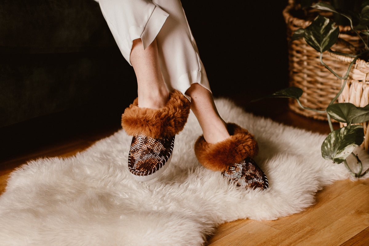 Women's feet wearing brown snake pattern sheepskin slippers, resting on a sheepskin rug.