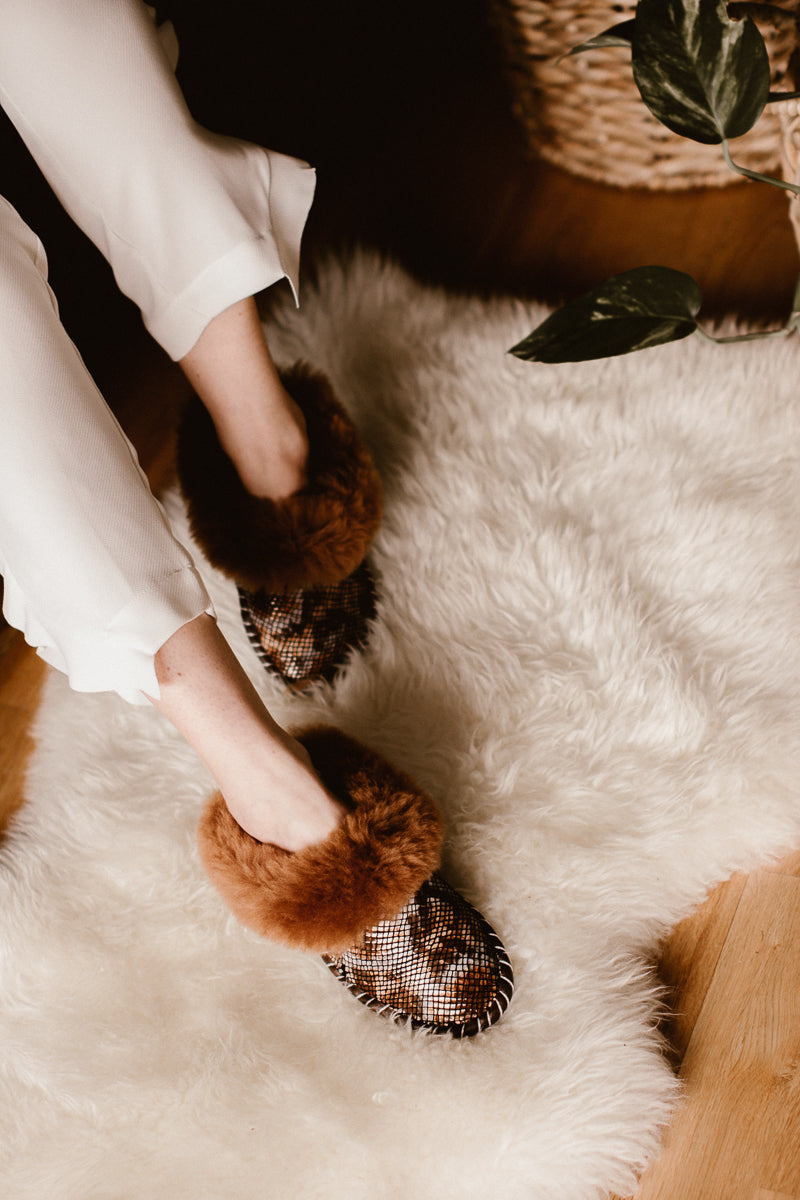 Women's feet wearing brown snake pattern sheepskin slippers, resting on a sheepskin rug. Bamboshe animal collection. 