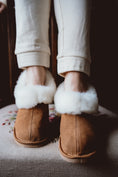 Load image into Gallery viewer, Classic sheepskin slippers with white fur, worn by a woman and placed on a hand-embroidered chair.

