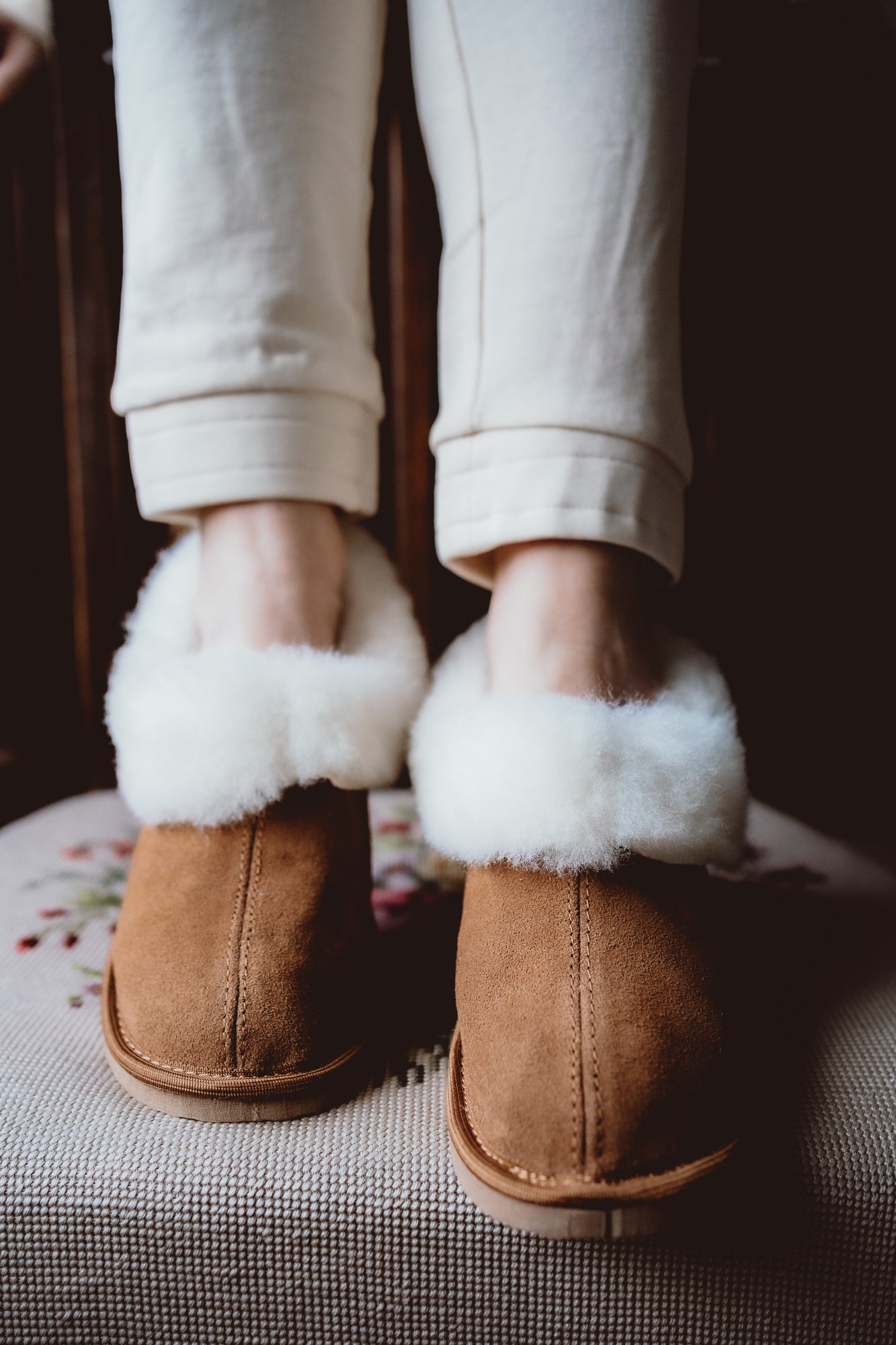 Classic sheepskin slippers with white fur, worn by a woman and placed on a hand-embroidered chair.